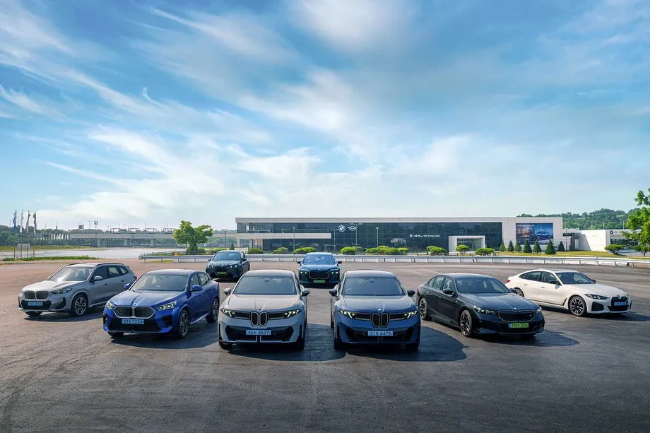 BMW 전동화 모델들이 BMW 드라이빙 센터 건물 앞 아스팔트 광장에 앞뒤 두 줄로 비스듬히 늘어서 있다. 흰색과 파란색, 회색 SUV와 세단이 섞여 있고 앞 범퍼 아래로 파란색 전기차 번호판이 보인다