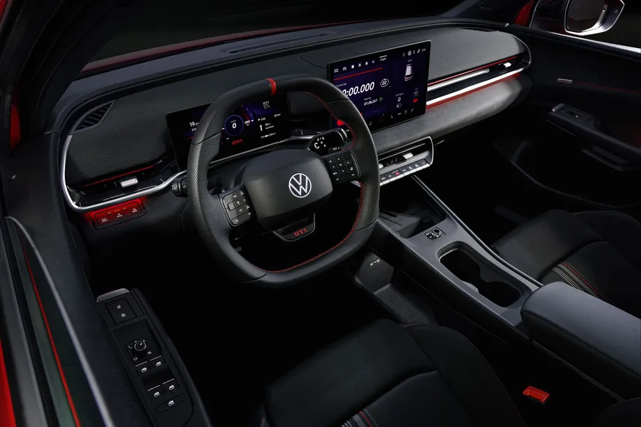 ID. Polo GTI 운전석 쪽 실내. 스포츠 스티어링 휠 12시 방향에 빨간 표식이 있고 가운데 아래에 빨간 GTI 글자가 보인다. 그 너머 디지털 계기판과 오른쪽 가로형 중앙 디스플레이에 랩타임 화면이 떠 있으며, 디스플레이 아래로 물리 버튼 줄이 가로로 놓여 있다